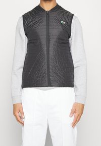 Svart kviltad väst med ståkrage och dragkedja fram. Har horisontell sömnad, en liten grön logotyp och är lager på en grå långärmad tröja.