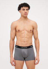 Sive bokserice z črnim pasom, ki nosi logo "LACOSTE", izdelane iz gladkega, raztegljivega blaga. Tesno prilegajoča zasnova.
