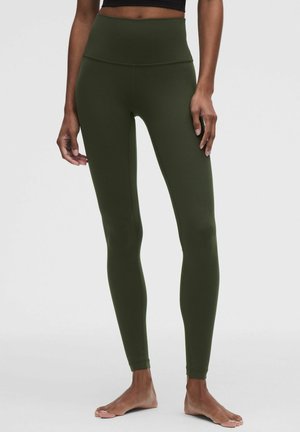 Kvinde iført olivengrønne leggings med høj talje, stående barfodet med arme afslappet ned langs siderne mod en ensfarvet baggrund.