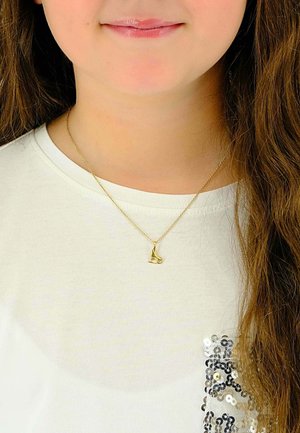 Collier en or avec un petit pendentif en forme de chaussure, porté sur une chemise blanche avec un accent en sequins sur le côté. De longs cheveux bruns ondulés sont visibles.