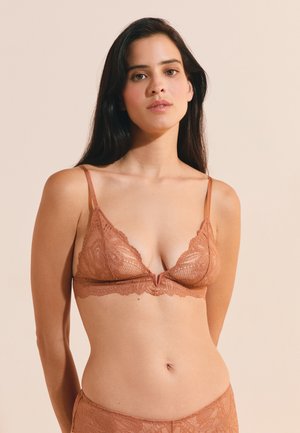 Donna con lunghi capelli scuri che indossa un reggiseno e biancheria intima in pizzo marrone coordinati, in piedi davanti a uno sfondo beige neutro.