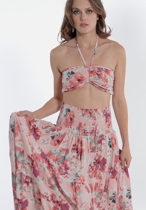 Donna che indossa un crop top a fiori rosa con collo halter e gonna lunga abbinata, tiene la gonna con una mano su uno sfondo semplice.