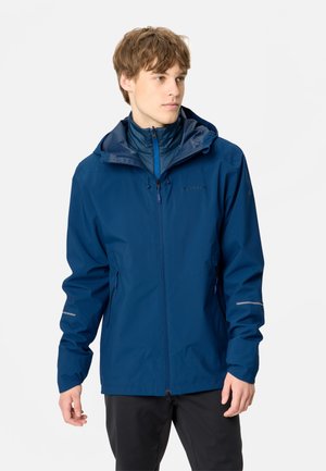 Jeune homme portant une veste à capuche bleue zippée, les mains détendues le long du corps, regardant vers la droite sur un fond clair uni.