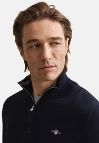 Mörkblå pullover med kvartszip, stickad struktur och en liten logga på bröstet. Kragen har kontrasterande randdetalj.