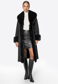 Dlouhý černý kabát s límcem a manžetami z umělé kožešiny, přes úzký bílý rolák a lesklou černou mini sukni, doplněný nadkolenkovými černými botami.