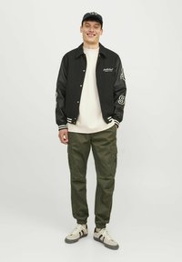 Chaqueta bomber negra con acentos blancos, sudadera beige, pantalones cargo verde oliva y zapatillas beige con rayas negras, posado frente a un fondo blanco.