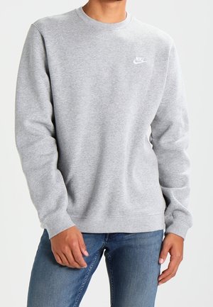 Mann trägt einen hellgrauen Nike-Sweatshirt mit kleinem weißem Logo auf der Brust und blauen Jeans, steht vor einem einfarbigen Hintergrund.