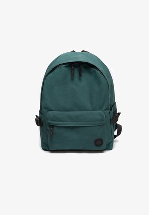 Zielony plecak z płótna o zaokrąglonym kształcie, z kieszenią na przodzie, dwoma czarnymi suwakami oraz okrągłym logo materiałowym na dolnej części przodu.