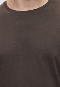 Nahaufnahme des oberen Brust- und Halsbereichs einer Person, die ein schlichtes dunkelbraunes Rundhals-Shirt trägt.
