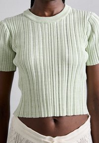 Osoba o ciemnej karnacji ubrana w jasnoróżowy prążkowany krótki top i kremowe dzianinowe spodnie, pokazana od szyi do talii.