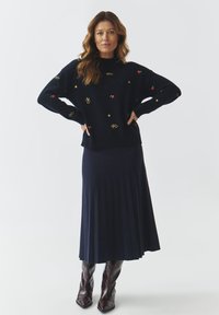 Maglione lavorato a maglia blu navy con ricami di ciliegie e stelle, abbinato a una gonna plissettata blu navy e stivali con tacco marroni, mostrato su uno sfondo bianco.