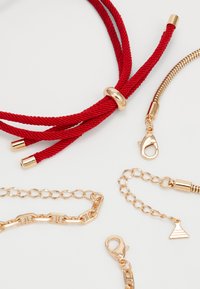 Collier en cordon rouge avec des accessoires en ton doré, comportant trois brins et une perle centrale. Inclut des chaînes dorées et des fermoirs de textures diverses.