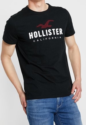 Man draagt een zwart Hollister California T-shirt en blauwe jeans, staat met één hand in zijn zak.