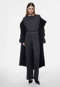 Czarna bluzka z długim rękawem w połączeniu z czarnym kombinezonem. Stylizacja jest uzupełniona obszerna czarną kurtką i zakończona spiczastymi czarnymi butami.