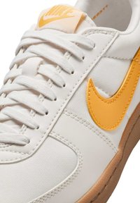 Valkoiset tennarit, joissa on oranssi Nike-logoswoosh, teksturoitu canvaspinta, kumipohja ja valkoiset nauhat. Näkyvät ompeleet korostavat muotoilulinjoja.