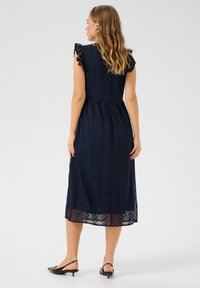 Marinblå spetsklänning med korta volangärmar. Har en figursydd bodice, midjesöm och en halvgenomskinlig nederkant med geometriska mönster.