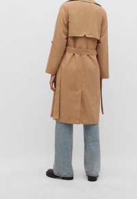 Kaki trenchcoat med bälte i midjan, med en ryggklaff och långa ärmar. Kombineras med vida ljusblå jeans och svarta stövlar.