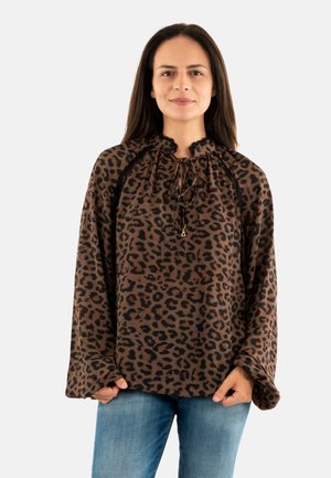 Femme aux longs cheveux bruns portant un blouse léopard brun et noir avec des détails en dentelle et un jean bleu clair, se tenant devant un fond blanc.