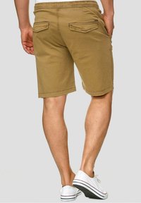 Homme portant un short beige jusqu'aux genoux et des baskets blanches, debout devant un fond uni gris clair.