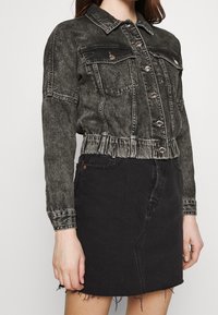 Žena v opraném černém jeanu s knoflíkovou přední stranou a náprsními kapsami, doplněná černou džínovou sukní s třepeným lemem.