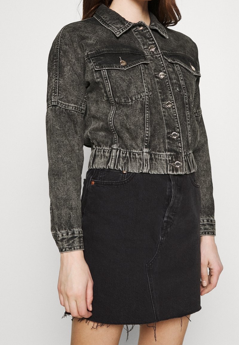 Žena v opraném černém jeanu s knoflíkovou přední stranou a náprsními kapsami, doplněná černou džínovou sukní s třepeným lemem.