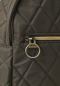 Sötétzöld, steppelt anyag, gyémántmintázattal a táska zsebében, kerek, réz cipzárhúzóval, amelyen a "Barbour" felirat található.