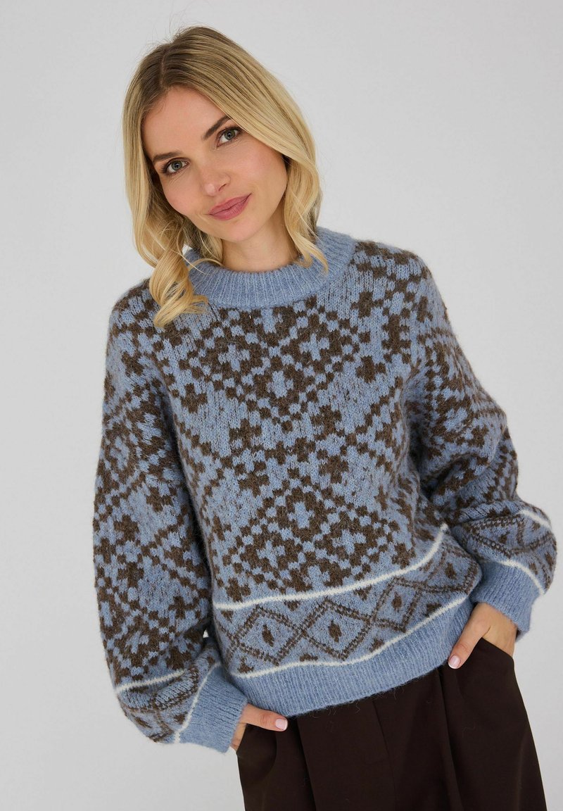 Femme blonde aux yeux bleus portant un pull en maille bleu et marron à motifs et un pantalon foncé, les mains dans les poches.