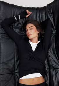 Schwarzes Longsleeve-Wickeloberteil mit tiefem V-Ausschnitt, über einem weißen Shirt getragen. Der Stoff ist glatt, und das Design hat eine figurbetonte Silhouette.