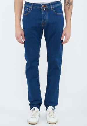 Vista frontale di una persona che indossa jeans blu aderenti con sneakers bianche e una maglietta bianca, in piedi con le braccia rilassate lungo i fianchi.