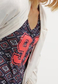 Niebieska koszulka na ramiączkach w wzory geometryczne w czerwieni i bieli, z dużą czerwoną cyfrą "91" nadrukowaną, nałożona pod jasnobeżowym kapturem.