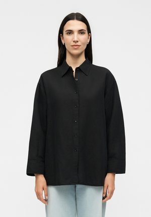Γυναίκα με ίσια σκούρα μαλλιά φοράει ένα φαρδύ μαύρο πουκάμισο με κουμπιά και ανοιχτομπλε τζιν, στέκεται μπροστά σε απλό λευκό φόντο.