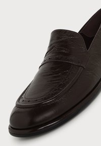 Gros plan sur un mocassin en cuir brun foncé avec bout arrondi, surpiqûres décoratives et détails perforés sur le dessus.