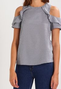 Bluzka w kratkę vichy z wycięciami na ramionach, marszczonymi rękawami i okrągłym dekoltem, wykonana z czarno-białej tkaniny.