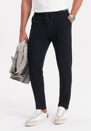 Pantalones negros ajustados con una tela texturizada, que cuentan con una cintura elástica con cordón y bolsillos laterales. Combinados con zapatillas blancas y una chaqueta gris claro.