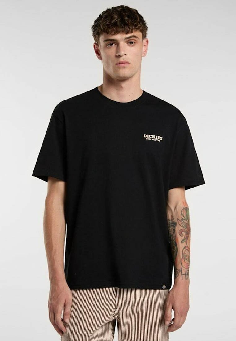 Ung mand med kort krøllet hår bærer en sort "Dickies Deep Roots" T-shirt og stribede bukser, og viser en farverig tatovering på sin venstre underarm.