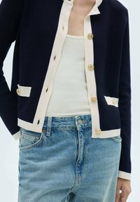 Cardigan bleu marine avec bordure crème et boutons ronds dorés, comprenant deux poches avant, porté sur un débardeur clair et un jean bleu déchiré.