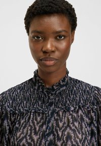 Camicia con bottoni realizzata in un tessuto scuro e fantasia, dotata di un colletto a balze e dettagli di arricciatura sulle spalle.