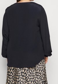Marineblauwe blouse met lange mouwen en een ronde halslijn, iets langere achterzoom en elastische gathered manchetten, gedragen over een bloemenpatroonrok.