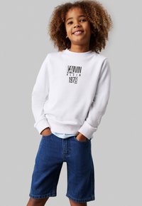 Felpa bianca con logo nero "CALVIN KLEIN 1978", abbinata a pantaloni corti di denim blu. Il modello ha i capelli ricci e le mani in tasca.