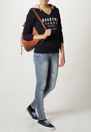 Person iført sort hættetrøje med hvid tekst, lyseblå slidte jeans, marineblå sneakers og bærer en brun læder skuldertaske.