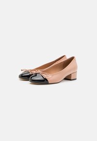 Chaussures en cuir verni beige avec un capuchon en cuir verni noir. Dotées d'un détail en nœud à l'avant et d'un petit talon en bloc.