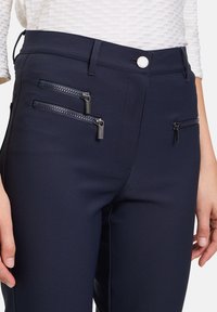 Mörkblå byxor med flera fickor med dragkedja och metallelement. Tillverkade av ett mjukt, stretchigt material. Knäppning med knapp i midjan.