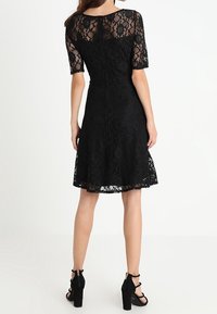 Robe en dentelle noire avec des manches jusqu'au coude, ourlet scallopé, corsage ajusté et jupe évasée. Associée à des escarpins à brides chevilles noirs.