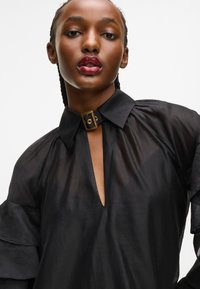 Μαύρη διαφανής μπλούζα με βαθύ ντε�κολτέ, δομημένους ώμους και φαρδιά μανίκια. Διαθέτει λεπτομέρεια κολάρου με χρυσό κούμπωμα. Λείος υφασμάτινος χαρακτήρας.