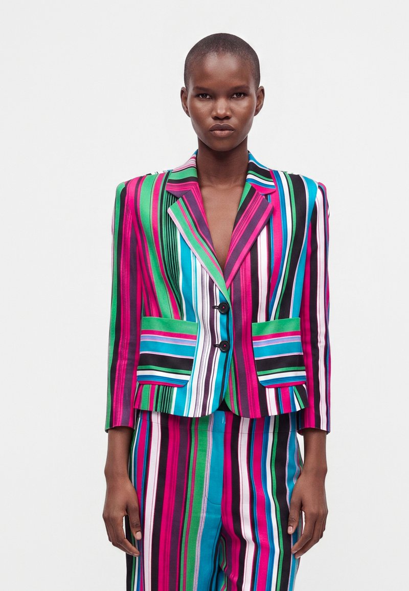 Femme portant un blazer rayé audacieux et coloré ainsi qu'un pantalon assorti en rose, vert, bleu, blanc et noir, debout devant un fond blanc.