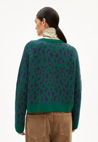 Grüner Strickpullover mit lila Leopardenmuster, knapper Schnitt, gerippte Bündchen und Saum. Wird über einem beigen Kragenhemd aus strukturiertem Stoff getragen.