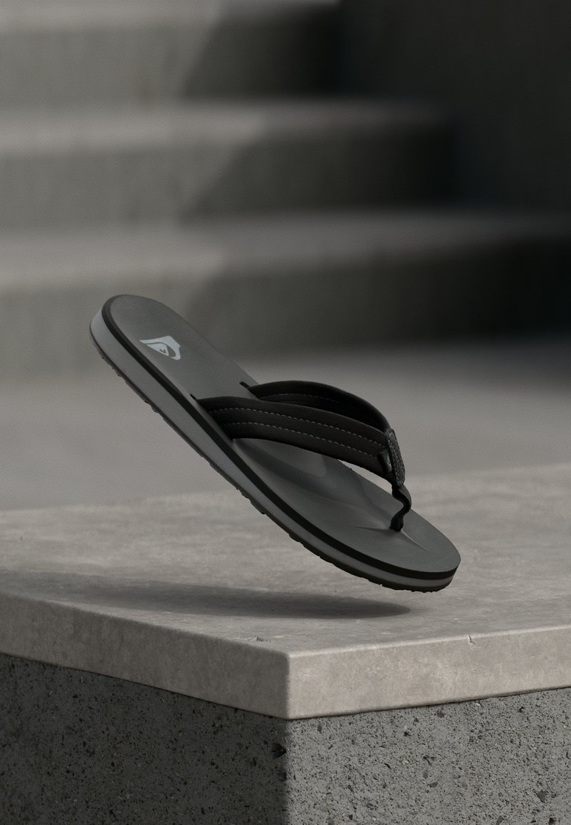 Μαύρη σαγιονάρα με ραμμένες λουρίδες αιωρείται πάνω από μια τσιμεντένια προεξοχή με θολή θέα σε σκαλοπάτια στο βάθος.