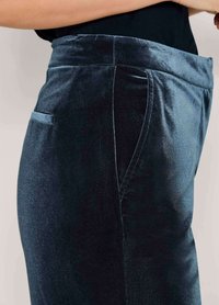 Close-up van donkerblauwe fluwelen broek gedragen door een persoon met hun arm op hun heup, waarbij de textuur van de stof en de zijzak worden benadrukt.