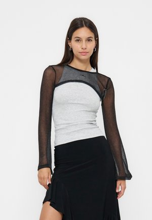 Femme aux longs cheveux noirs portant un haut gris moulant superposé à un haut en résille noire transparente à manches longues et une jupe noire avec un volant sur le côté.