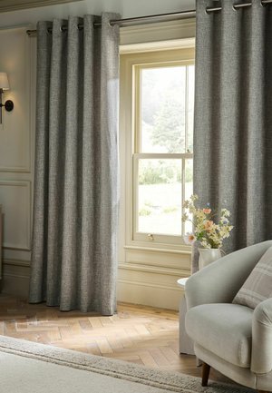 Gray textured curtains hang partially open on a window in a bright room with a light armchair and floral arrangement on a side table.